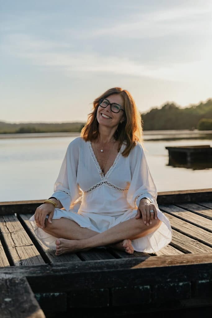 Femme souriante aux cheveux roux et lunettes, en robe blanche, assise sereinement sur une jetée en bois au bord d'un lac au coucher du soleil.