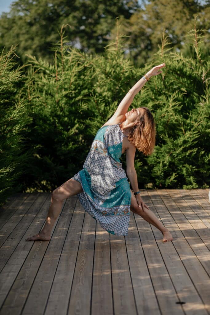 Femme pratiquant le yoga en extérieur sur un ponton en bois, bras tendus dans une pose latérale. Elle porte une robe turquoise et est entourée de verdure luxuriante.