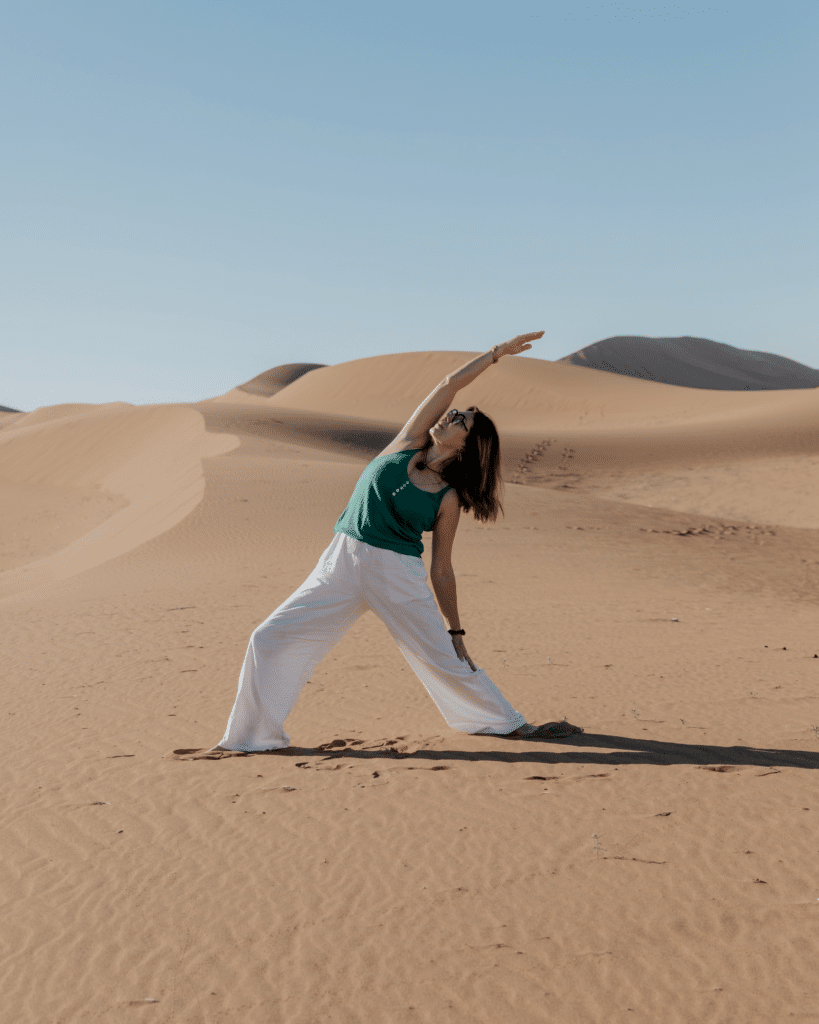 Femme en yoga étirement dans les dunes du désert, haut vert et pantalon blanc, ciel bleu dégagé.