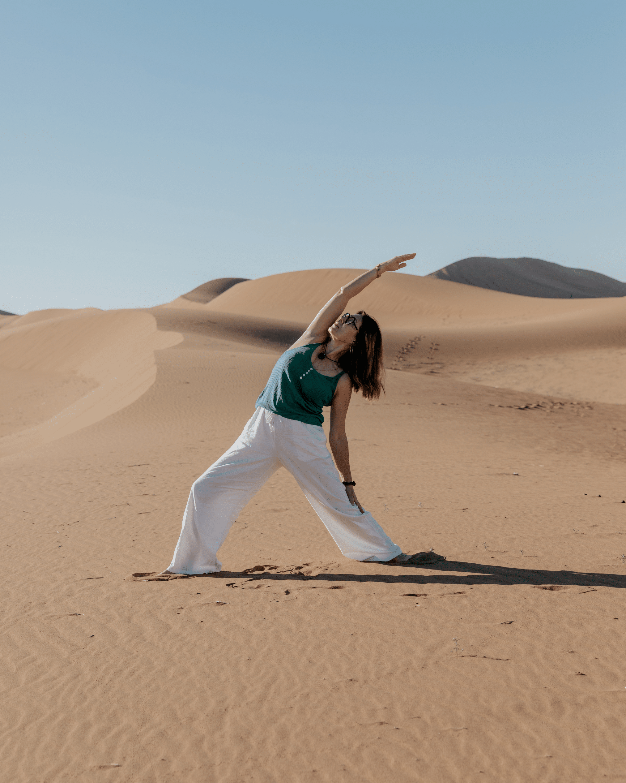 Femme en yoga étirement dans les dunes du désert, haut vert et pantalon blanc, ciel bleu dégagé.