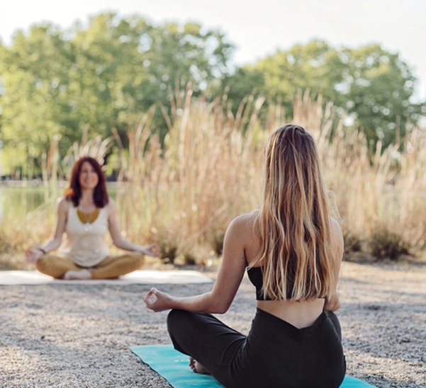 Deux femmes font du yoga assis en extérieur. L'une est de dos, l'autre face. Scène paisible au bord de l'eau.