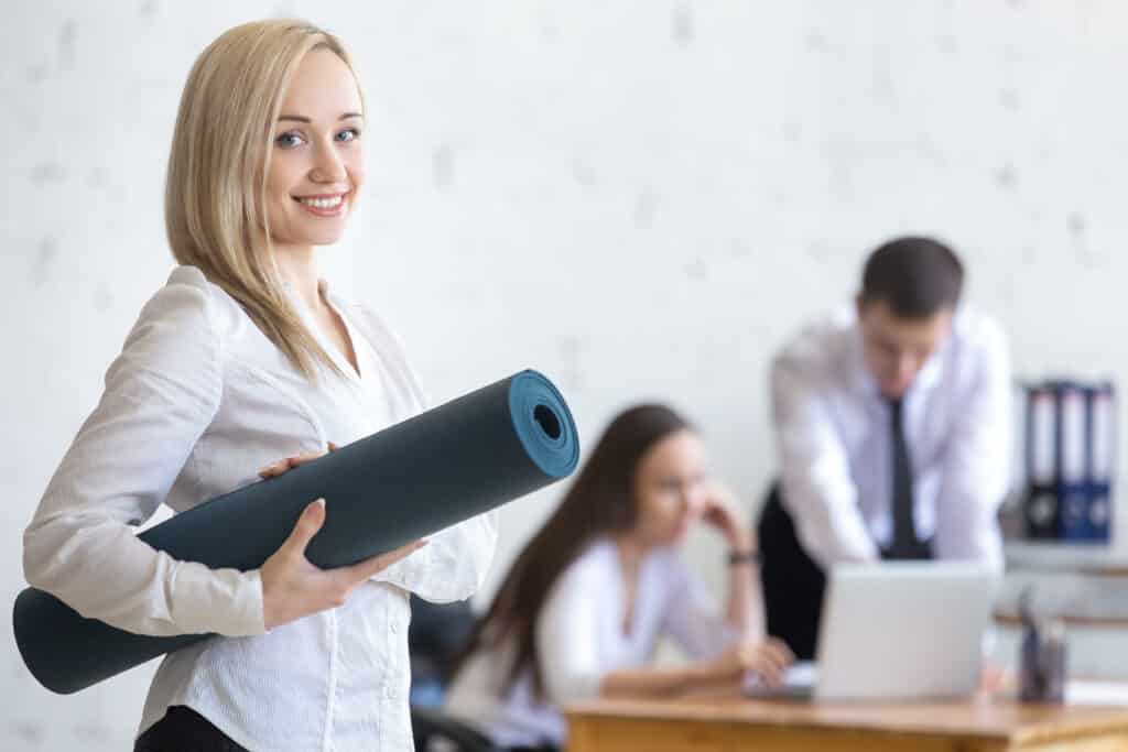 Femme blonde souriante tenant un tapis de yoga bleu dans un bureau moderne avec des collègues flous en arrière-plan, symbolisant le bien-être au travail.