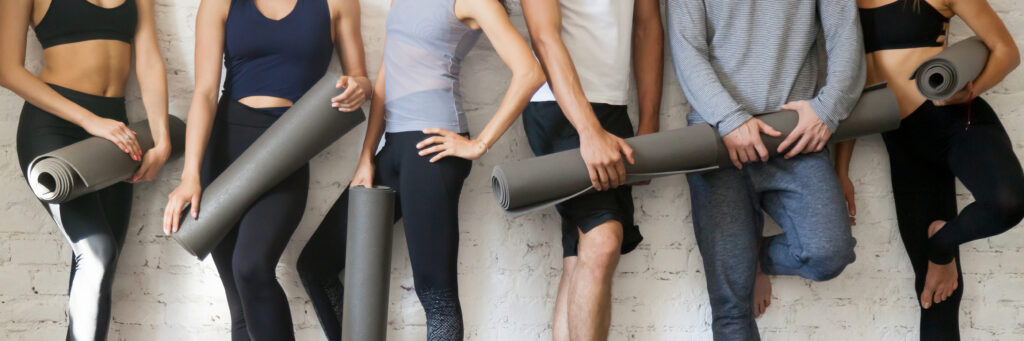 Groupe de personnes diverses en tenue de sport, tenant des tapis de yoga roulés contre un mur blanc, prêtes pour l'entraînement.