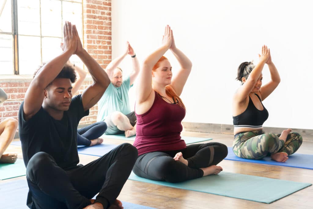Groupe diversifié d'adultes méditant au yoga, assis sur des tapis, mains jointes au-dessus de la tête dans un studio lumineux.