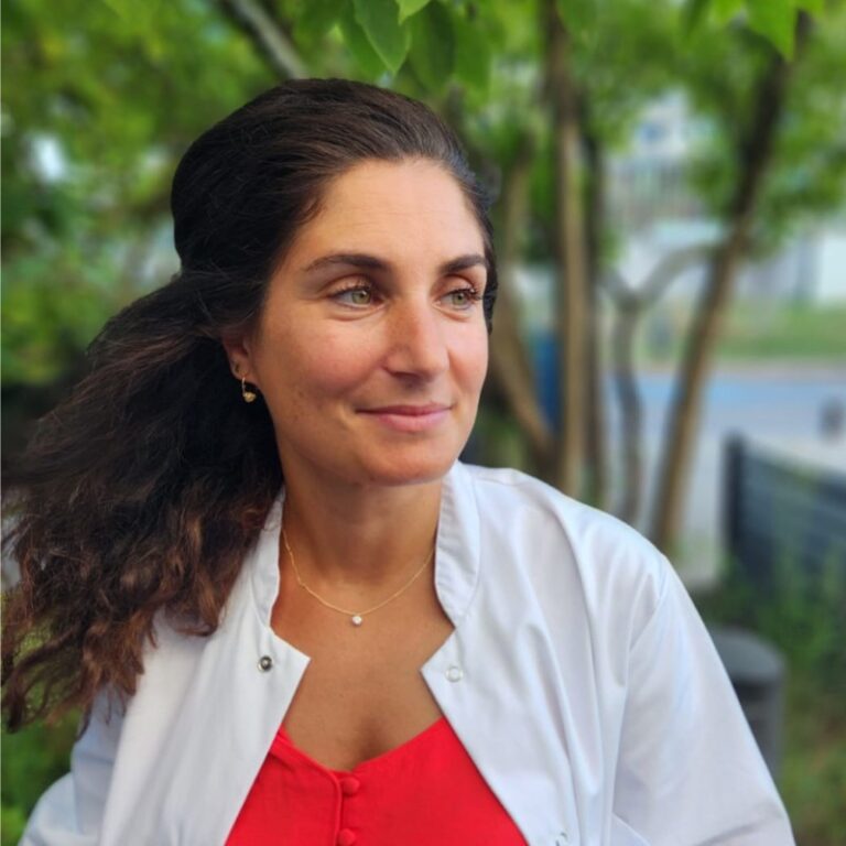 Femme professionnelle souriante en blouse blanche et haut rouge, collier doré. Cheveux bruns, yeux verts, regard doux, fond extérieur flou verdoyant.