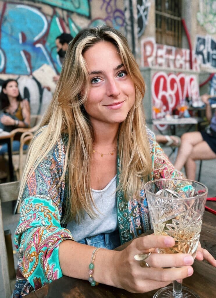 Sourire bohème et verre frais sur fond urbain Jeune femme souriante aux cheveux blonds, vêtue d'un kimono cachemire coloré, tenant un verre de vin blanc devant un mur de graffitis.