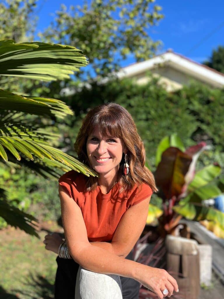 Portrait extérieur d'une femme souriante en t-shirt terracotta. Elle pose les bras croisés dans un jardin tropical ensoleillé. Bijoux ethniques visibles.
